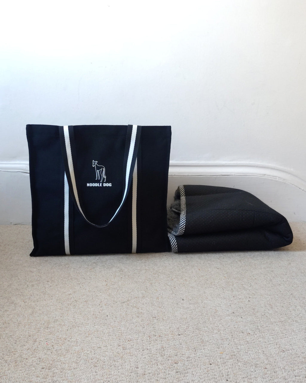 A black canvas tote bag with black and white handles and a white dog silhouette logo that reads 'NOODLE DOG' sits on a beige carpet next to a folded black mat against a white wall.