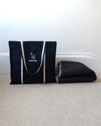 A black canvas tote bag with black and white handles and a white dog silhouette logo that reads 'NOODLE DOG' sits on a beige carpet next to a folded black mat against a white wall.