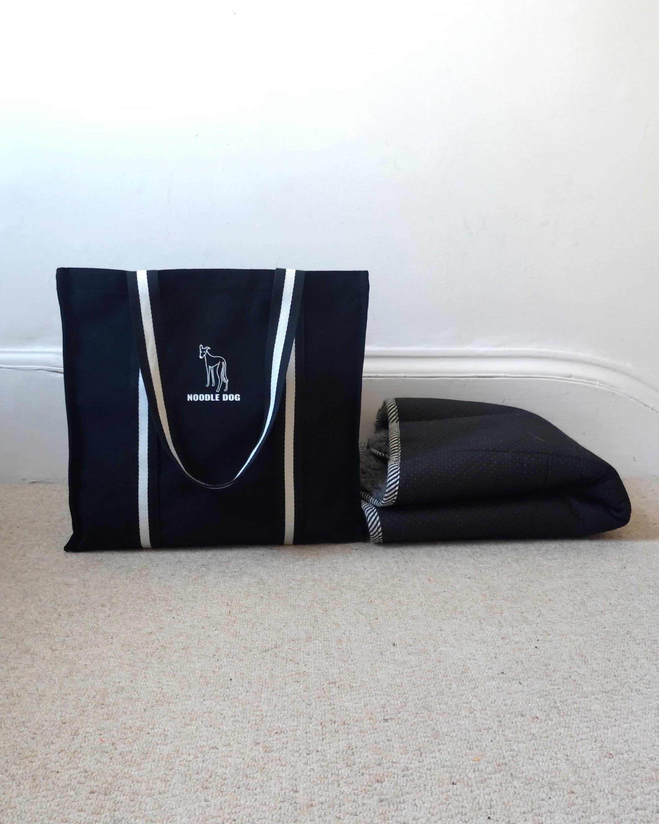 A black canvas tote bag with black and white handles and a white dog silhouette logo that reads 'NOODLE DOG' sits on a beige carpet next to a folded black mat against a white wall.
