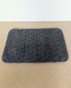 top view of a grey wool travel mat with black and white strip binding on a beige carpet and white wall. 