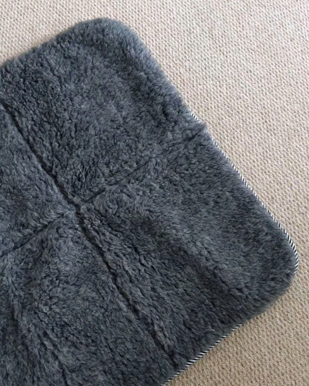 A close-up of a dark grey plush wool mat, showing its soft texture and stitched construction, placed on a beige carpeted floor.